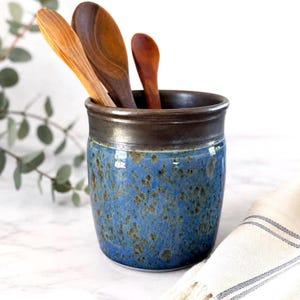 May include: A cylindrical ceramic utensil holder with a glossy blue glaze featuring dark brown speckled patterns and a solid dark brown rim. Three wooden spoons of varying sizes and wood tones are placed inside the container.