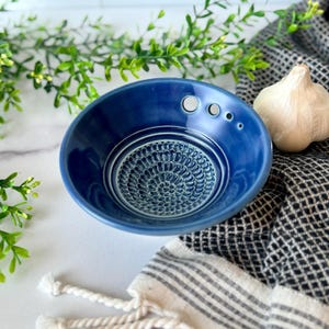 May include: A deep blue ceramic garlic grater bowl with a textured grating surface and three small holes. A bulb of garlic rests on a black and white patterned cloth, with greenery in the background.