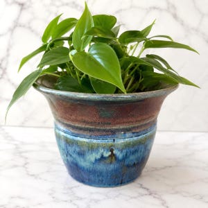May include: A ceramic planter with a flared rim and a glossy glaze featuring a horizontal band of deep brown at the top, transitioning into a vibrant blend of blue and teal hues at the base, containing a lush green heart-leaf philodendron plant.