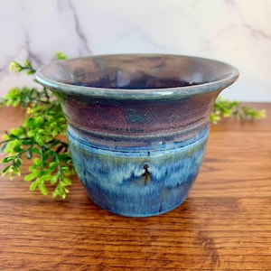 May include: A handcrafted ceramic vase with a wide, flared rim. The vase features a gradient of colors, with a deep blue base transitioning to a band of maroon and a dark teal rim. The pottery is glazed, giving it a glossy finish.