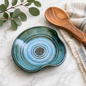May include: A round ceramic spoon rest featuring a glossy blue and green glaze with concentric circular grooves and a textured dark center medallion. The dish has a curved indentation on one side for a spoon handle and a smooth, glazed finish.