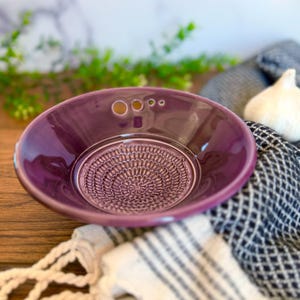 May include: A deep purple ceramic garlic grater bowl with a textured grating surface. The bowl has a round shape with a small handle and three holes. A bulb of garlic rests nearby on a patterned cloth.