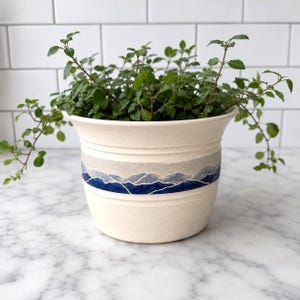 May include: A cream-colored ceramic planter with a flared rim and two horizontal ridges. A band encircles the center, featuring a painted landscape of layered blue and white mountains. A leafy green plant grows out of the top of the pot.