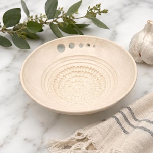 May include: A circular, off-white ceramic grater bowl featuring a textured, radial pattern at the center for grating and a series of five small, graduated circular holes along the upper rim for hanging or storage purposes.