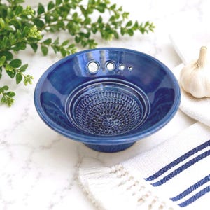 May include: A deep blue ceramic grater bowl with a textured, floral-patterned center for grating garlic or ginger. The bowl features a smooth, glossy finish and a series of four circular holes of varying sizes along the inner rim for herb stripping.