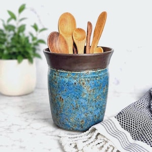May include: A cylindrical ceramic utensil crock featuring a speckled blue and brown glaze, topped with a dark brown rim. The container is filled with various light-colored wooden spoons and kitchen utensils of different shapes and sizes.