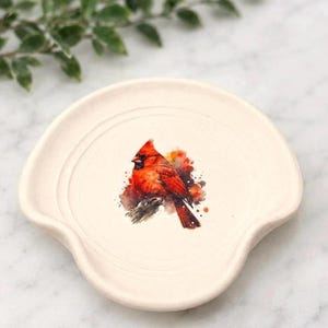 May include: A cream-colored ceramic spoon rest featuring a watercolor-style illustration of a bright red cardinal bird perched on a branch. The spoon rest has a circular center with a raised rim and a curved indentation for the spoon handle.