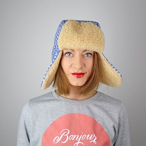 May include: A woman wearing a blue and white patterned trapper hat with a faux fur trim. The hat has ear flaps that are folded up. She is wearing a gray sweatshirt with a red circle and the word "Bonjour" written in white.
