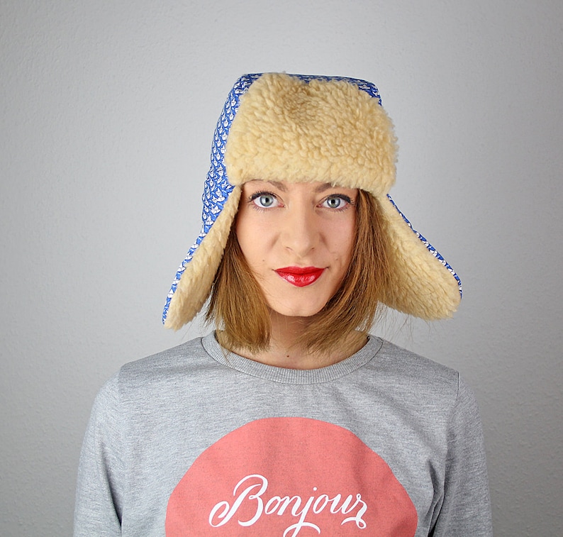 May include: A woman wearing a blue and white patterned trapper hat with a faux fur trim. The hat has ear flaps that are folded up. She is wearing a gray sweatshirt with a red circle and the word "Bonjour" written in white.