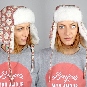May include: A pink and white patterned trapper hat with faux fur ear flaps and a white faux fur brim. The hat has a long tie that is tied under the chin. The hat is being worn by a person with short brown hair. The person is wearing a grey sweatshirt with a red circle and the text "Bonjour Mon Amour" in white.