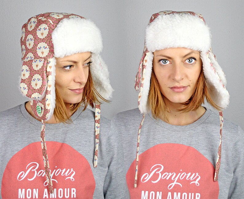 May include: A pink and white patterned trapper hat with faux fur ear flaps and a white faux fur brim. The hat has a long tie that is tied under the chin. The hat is being worn by a person with short brown hair. The person is wearing a grey sweatshirt with a red circle and the text "Bonjour Mon Amour" in white.