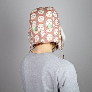 May include: A pink and white patterned winter hat with a fluffy white brim and green buttons. The hat is worn by a person with brown hair.