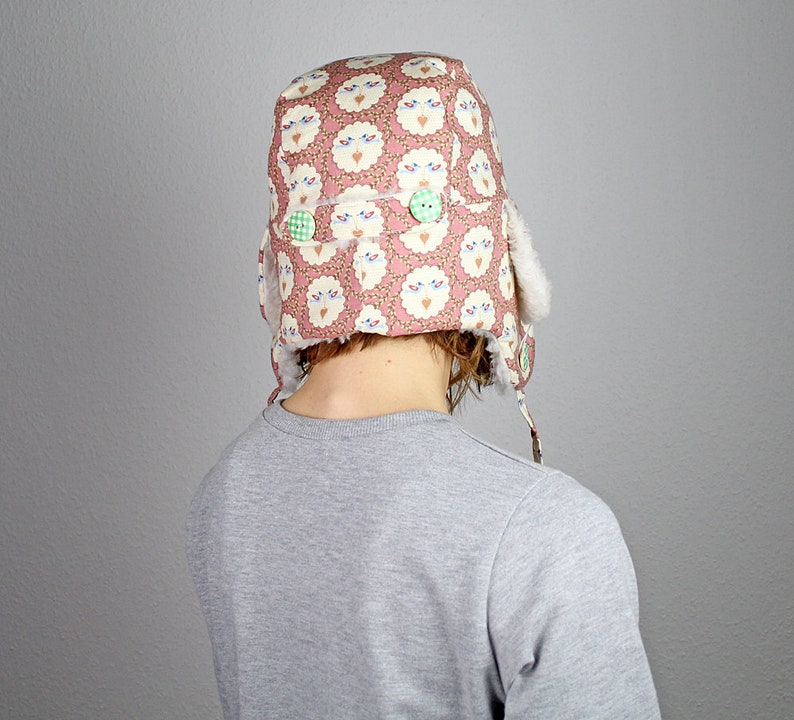 May include: A pink and white patterned winter hat with a fluffy white brim and green buttons. The hat is worn by a person with brown hair.