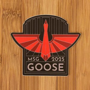 May include: A sticker with a stylized red goose design, featuring outstretched wings and a central arrow shape, set against a black background. The text "MSG 2025 GOOSE" is displayed below the design in a rectangular frame.