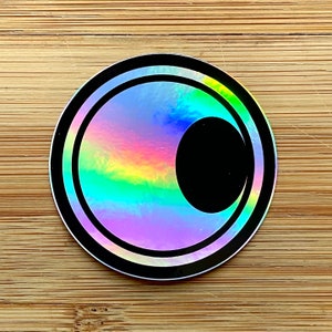 May include: A holographic sticker with a black circle inside a double black circle. The sticker has a rainbow iridescent effect.