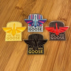 May include: Four enamel pins with a goose design and the text "MSG 2025 GOOSE". The pins feature a variety of color schemes, including yellow, blue, red, and black. The pins are set against a wooden background.
