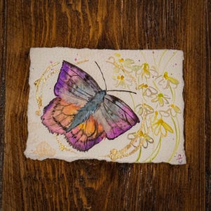 May include: Watercolor painting of a butterfly with purple, orange, and blue wings. The butterfly is surrounded by gold accents and small white and yellow flowers. The artwork is on textured paper.