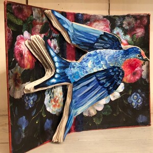 May include: A blue bird with red accents is depicted in a pop-up book with a floral background. The bird is made of paper and painted in a realistic style.