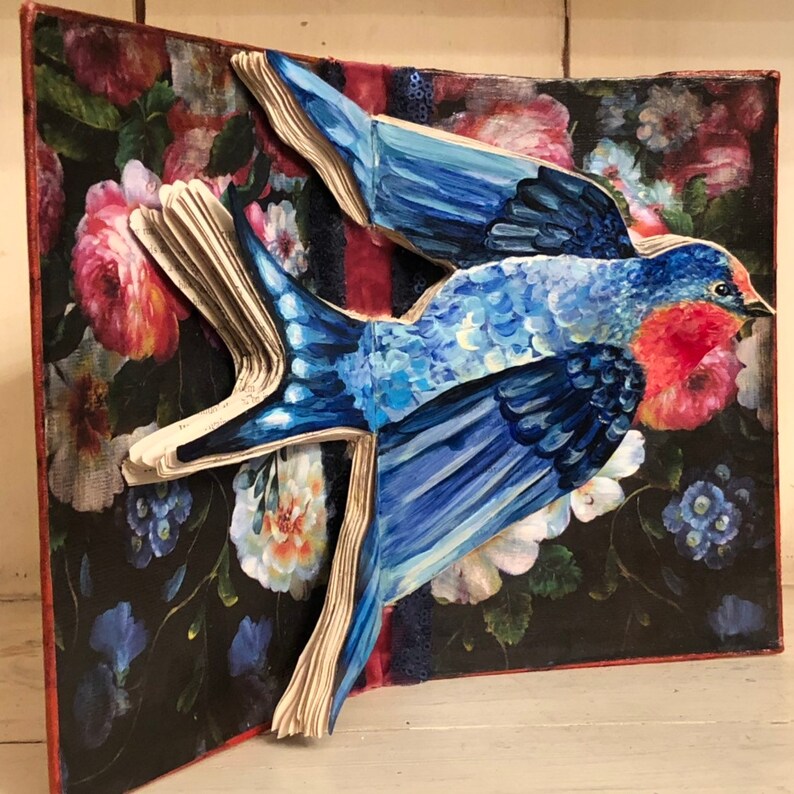 May include: A blue bird with red accents is depicted in a pop-up book with a floral background. The bird is made of paper and painted in a realistic style.