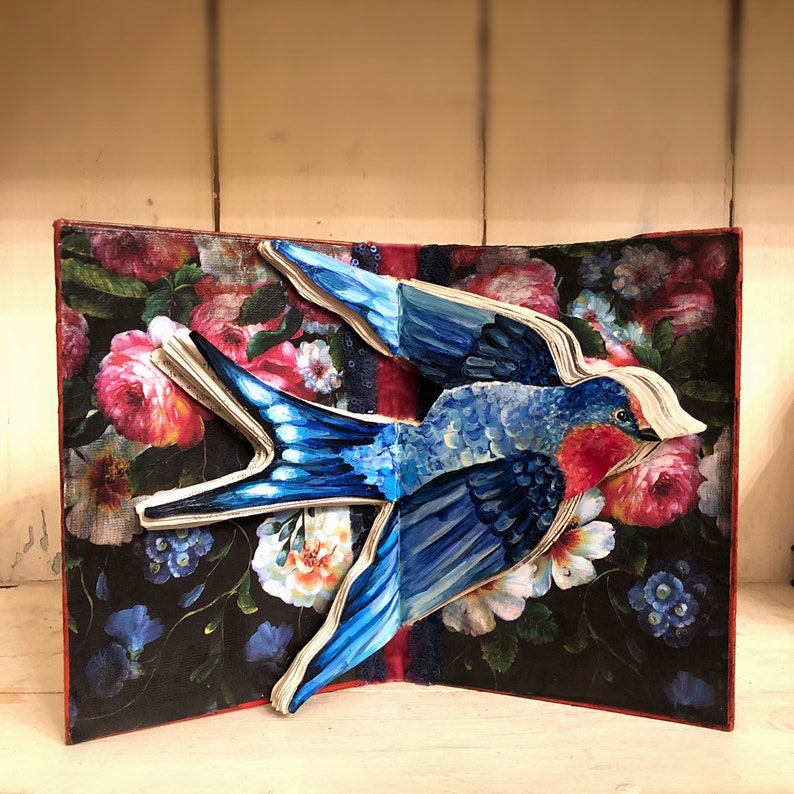 May include: A blue and red paper bird sculpture with a floral background. The bird is in flight and has a long tail. The background is a dark blue with red, pink, and white flowers.