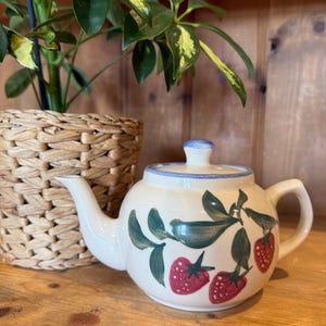 May include: A white ceramic teapot decorated with strawberries. The teapot has red strawberries, green leaves, and a blue rim and lid. A woven basket and a potted plant are in the background.