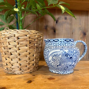 May include: A blue and white ceramic jug with a detailed bird and floral pattern. The jug has a curved handle and a decorative rim. Next to it is a woven basket planter with a green plant.