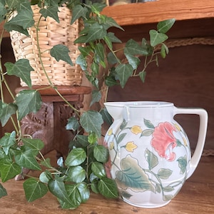 May include: A white ceramic jug featuring a painted floral pattern with large coral-coloured blossoms, green leaves, and small yellow buds. The jug has a rounded body, a wide spout, and a sturdy handle, with a smooth, glossy finish.