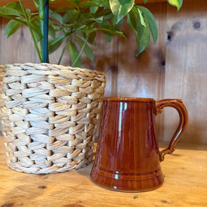 May include: A brown ceramic mug with a handle, placed on a wooden surface. The mug features vertical ridges and a glossy finish. A woven basket and a green plant are in the background.