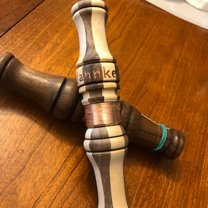 Könnte beinhalten: Zwei hölzerne Entenrufe mit einem gestreiften Design. Der obere Ruf hat ein Kupferband und den Namen "Jahnke" auf der Seite eingraviert.