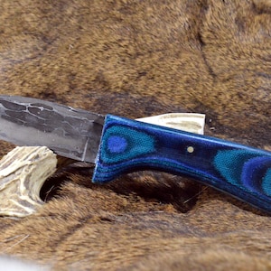 May include: A handcrafted knife with a textured blade and a vibrant blue and teal handle. The handle features a swirling pattern and is secured with a single silver pin. A small piece of antler rests near the blade.