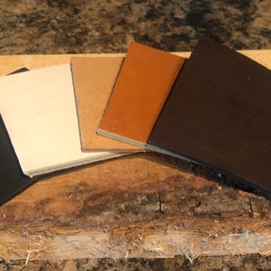 Puede incluir: Cinco muestras de cuero en diferentes colores: negro, blanco, marrón, naranja y marrón oscuro. Las muestras están dispuestas en forma de abanico sobre una superficie de madera.