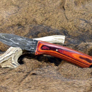 May include: A handmade knife with a patterned blade and a wooden handle in shades of red and orange. The knife rests on a brown, textured surface, with a piece of antler supporting the blade.