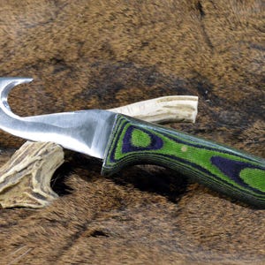 May include: A hunting knife with a curved blade and a green and purple patterned handle. The knife is resting on a brown, textured surface, with pieces of antler near the blade.
