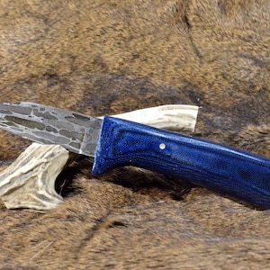 May include: A handcrafted knife with a patterned blade and a vibrant blue handle. The knife rests on a piece of antler against a backdrop of brown fur, showcasing its rustic aesthetic.