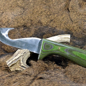 May include: A hunting knife with a curved blade and a green and black wooden handle. The knife is resting on a brown, textured surface, with a piece of antler near the handle.
