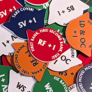 Puede incluir: Una colección de fichas de juego coloridas con varios textos y símbolos. Las fichas son en tonos de rojo, naranja, azul, verde, blanco y negro. El texto incluye "Take Cover!", "First Rank, Fire! Second Rank, Fire!" y valores numéricos.
