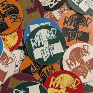 May include: A collection of circular coasters featuring a variety of solid background colours including orange, blue, dark green, red, and white. Each coaster displays the text RILED UP in a stylised, distressed white or dark font across the centre.