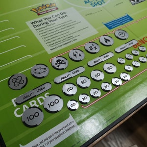 May include: Green Pok&eacute;mon Trading Card Game playmat with instructions and game markers. The playmat features the Pok&eacute;mon logo and text, along with various silver game tokens with symbols and numbers.