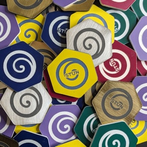 May include: A selection of hexagonal tokens in diverse colours, such as blue, yellow, purple, and green. Each token displays a spiral design or the word "STUN" in a circular pattern. The tokens have a metallic or brushed finish.