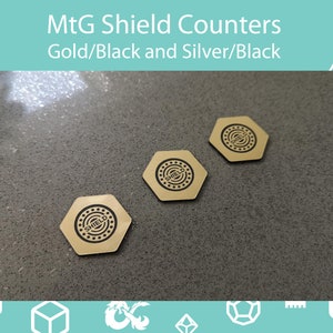 Op de afbeelding: Drie goudkleurige, zeshoekige spelcounters met zwarte tekst en een cirkelvormig ontwerp. De counters zijn gelabeld met "SHIELD" en zijn bedoeld voor gebruik in het spel Magic: The Gathering. De tekst "MtG Shield Counters Gold/Black and Silver/Black" staat bovenaan.