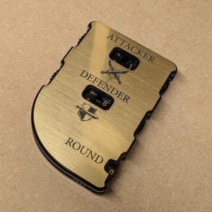 Op de afbeelding: Een goudkleurig multi-tool met zwarte rand. De tool heeft de woorden "ATTACKER", "DEFENDER" en "ROUND" gegraveerd. Het heeft twee tellers met cijfers en gekruiste zwaarden en schildafbeeldingen.