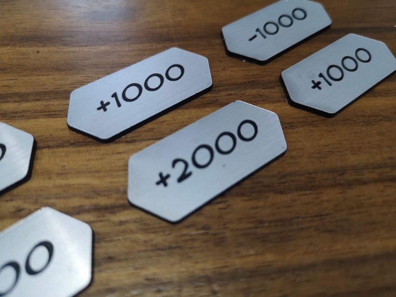 K&ouml;nnte beinhalten: Silberfarbene Metall-Token mit schwarzem Text, der Werte von +1000, -1000 und +2000 angibt. Die Token haben eine einzigartige Form mit schwarzem Rand und liegen auf einer Holzoberfl&auml;che.