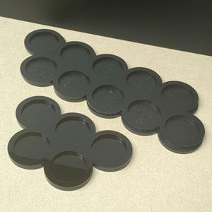 Peut inclure: Deux plateaux noirs en forme de fleur avec des empreintes circulaires. L'un des plateaux comporte six empreintes, l'autre dix. Les plateaux sont faits d'un mat&eacute;riau lisse et sombre, con&ccedil;us pour contenir de petits objets.