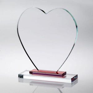 May include: A clear glass heart-shaped ornament or award. The heart sits on a rectangular base with a red accent. The base is made of clear glass, and the heart is outlined in black.