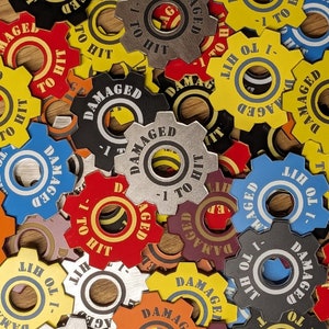 May include: A selection of brightly coloured, gear-shaped tokens, each bearing the words "DAMAGED" and "-1 TO HIT". The tokens, in shades like yellow, red, blue, and black, are scattered across a wooden surface.