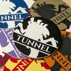 Puede incluir: Una colección de fichas circulares de colores con la palabra "TUNNEL" impresa. Las fichas presentan un gráfico estilizado y están en varios colores, incluyendo azul, negro, blanco y naranja. Las fichas están dispersas.