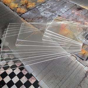 Puede incluir: Una pila de láminas acrílicas rectangulares transparentes. Las láminas están abanicadas, revelando su transparencia. El fondo presenta una superficie texturizada de temática industrial con una mezcla de patrones grises, marrones y negros.