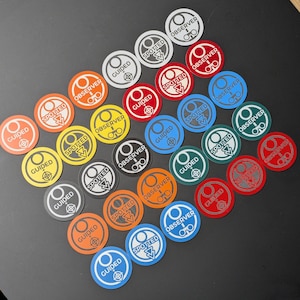 May include: A collection of colorful, circular game tokens with various designs and text. Each token features the words "GUIDED", "SPOTTED", or "OBSERVER", along with a graphic. The tokens are in shades of orange, red, blue, green, yellow, and gray.