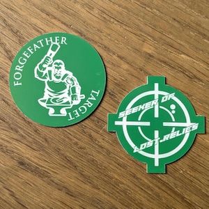May include: Two green stickers on a wooden surface. One is round with a white illustration of a blacksmith and the text "FORGEFATHER TARGET". The other is a crosshair shape with the text "SEEKER OF LOST RELICS".