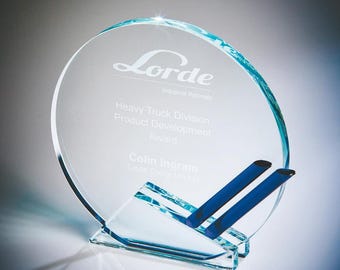 Może przedstawiać: Przezroczysta szklana nagroda o okrągłym kształcie, z tekstem "Lorde Industrial Materials" i "Heavy Truck Division Product Development Award". Nagroda ma niebieskie akcenty i podstawkę.